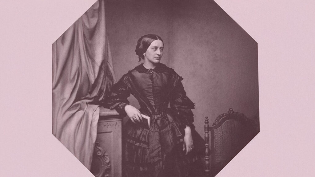 Stående Clara Schumann bär mörk klänning och lutar höger arm mot en byrå. Allvarsamt blickar hon åt sidan.